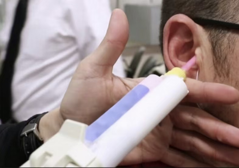 A close-up image of a person having an ear impression taken with an impression gun, held by a professional in a clinical setting, with another individual in a white shirt and tie in the background