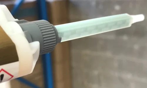 Close-up of a dual-cartridge caulking gun equipped with a long, translucent static mixing nozzle for dispensing and automatically mixing two-component epoxy adhesive. The nozzle is attached to the cartridge, ready for application in a workshop setting